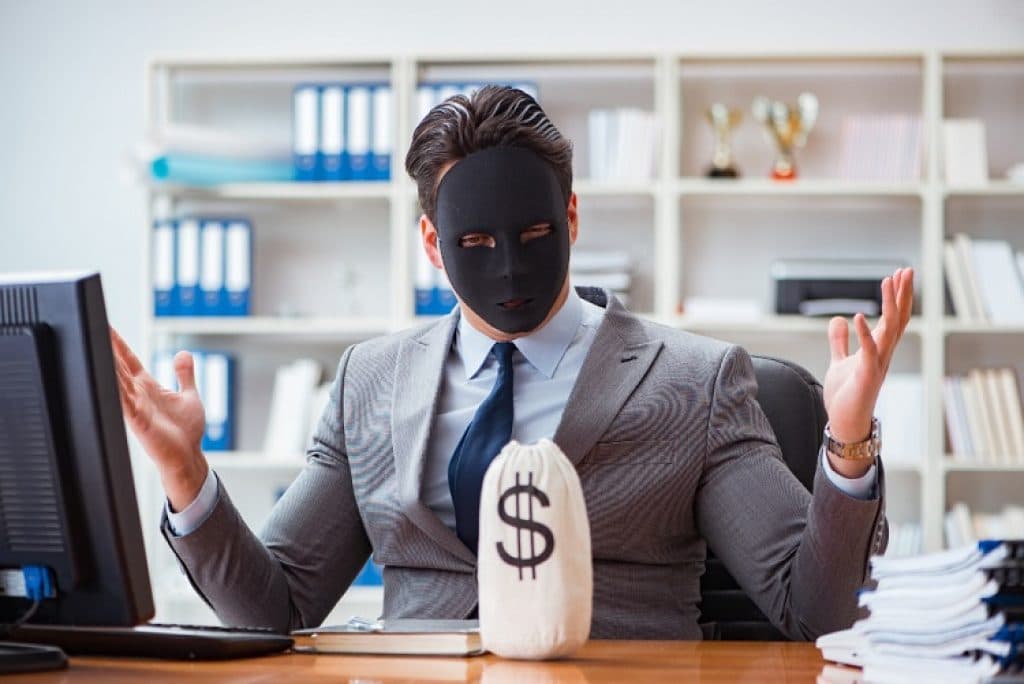 גבר עם מסיכה שחורה יושב בחליפה מול שולחן עם שק של כסף